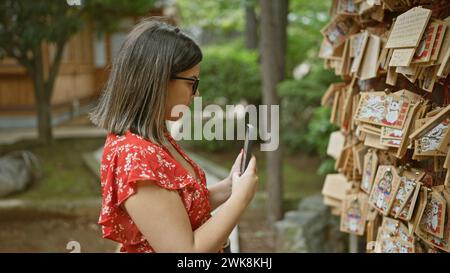 Wunderschöne hispanische Frau in Brille, die Gebetstafeln im japanischen Gotokuji-Tempel festnimmt und die Tradition durch ein Smartphone-Objektiv bewahrt Stockfoto