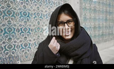Lächelnde junge Frau mit Brille und Schal vor einem kunstvollen Fliesenhintergrund im topkapi-Palast in istanbul Stockfoto