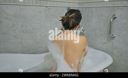 Rückansicht einer jungen Frau, die zu Hause ein Schaumbad in einem Marmorbad genießt. Stockfoto