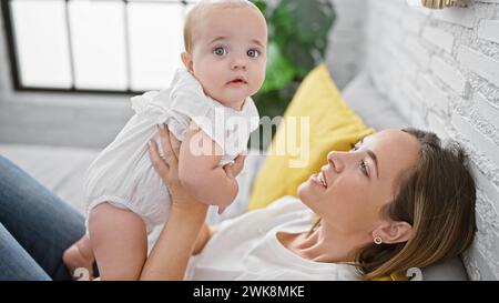 Fröhliche Mutter, die auf dem Bett liegt, selbstbewusst die glückliche Tochter in der Luft hält, Spaß hat, während sie lächelt und zusammen in einem warmen, gemütlichen Schlafzimmer spielt. Stockfoto