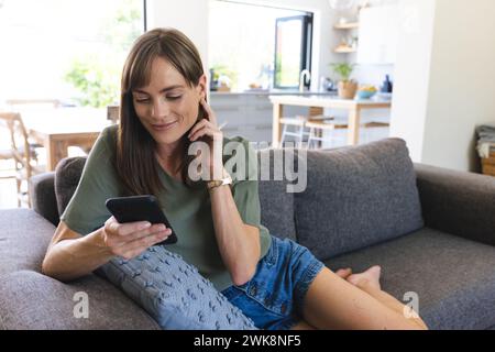Eine weiße Frau mittleren Alters entspannt sich auf einem Sofa, während sie ihr Smartphone durchsucht, mit Kopierraum Stockfoto