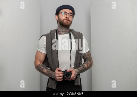 Cooler stylischer Mann professioneller Fotograf mit Brille im Bart mit Tattoo in Mode Strickmütze und Pullover hält Ein modernes spiegelloses Foto Stockfoto