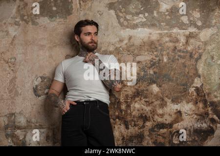 Ein hübscher, brutaler Hipster-Mann mit Frisur und Bart in Einem weißen T-Shirt mit Pullover steht neben einer alten Betonmauer Stockfoto
