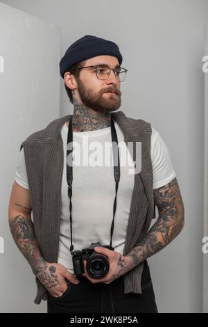 Modischer junger Mann Hipster-Fotograf mit Brille und Strickmütze in T-Shirt mit Pullover hält Eine moderne spiegellose Kamera und einen Ständer Stockfoto