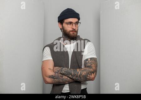 Coole, hübsche Hipster-Mann mit Bart, Schnurrbart und Tattoo mit stylischer Brille, die Einen Strickmütze in Einem modischen weißen T-Shirt mit Einem Pullover in Th trägt Stockfoto