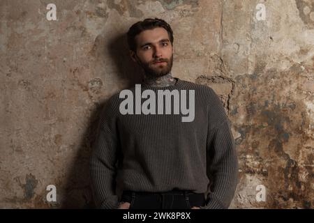 Das hübsche junge Mann Hipster Model mit Schnurrbart, Bart und Frisur in Einem modischen Strickpullover steht in der Nähe Einer Grunge Betonwand Stockfoto