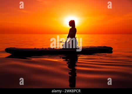 Frau, die sich auf dem Paddelbrett am Meer mit warmem, hellem Sonnenuntergang oder Sonnenaufgang entspannt. Stockfoto