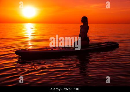 Silhouette einer Frau, die sich auf dem Paddelbrett am ruhigen Meer mit warmem, hellem Sonnenuntergang oder Sonnenaufgang entspannt. Stockfoto