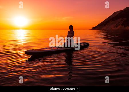 Schlanke junge Frau, die sich auf dem Paddelbrett am Meer mit warmem, hellem Sonnenuntergang oder Sonnenaufgang entspannt. Stockfoto