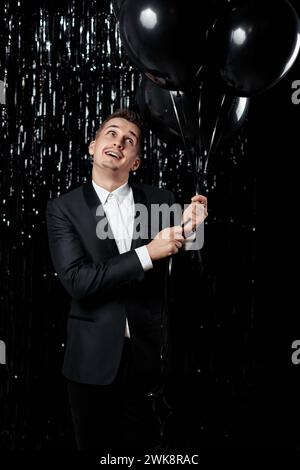 Glücklicher, gutaussehender Mann in schwarzer Jacke, der schwarze Luftballons auf Glitzerhintergrund hält. Stockfoto