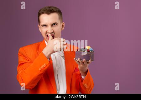 Hübscher Geburtstagsmann in orangefarbener Jacke, der Kuchen auf violettem Hintergrund isst Stockfoto