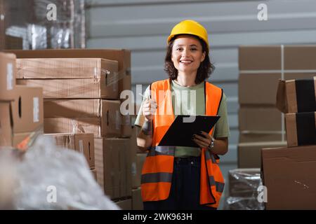 Fröhliche Lagerarbeiterin in Sicherheitsweste und Schutzhelm, die Klemmbrett und Stift in der Nähe von Fracht hält Stockfoto