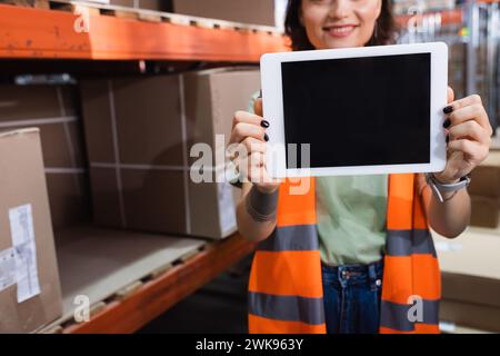 Beschnittene und glückliche Lagerarbeiterin in einer Sicherheitsweste, die ein digitales Tablet mit leerem Bildschirm hält Stockfoto