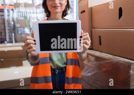 Abgeschnittene und lächelnde Lagerarbeiterin in Sicherheitsweste mit digitalem Tablet und leerem Bildschirm Stockfoto