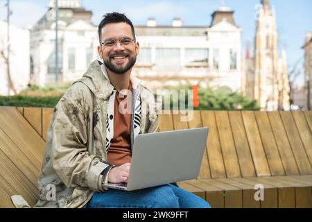 Junger Hipster Millennial Mann Weltreisender nomadischer Lifestyle im Freien sitzend mit Laptop Computer auf der Suche nach neuen Reiseziel und Pl Stockfoto