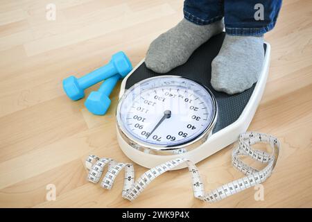 Füße eines Mannes in grauen Socken, die auf einer Waage stehen, um das Gewicht nach dem Fitnesstraining zu überprüfen, blaue Kurzhanteln und ein Maßband, das in der Nähe auf einem Wald liegt Stockfoto