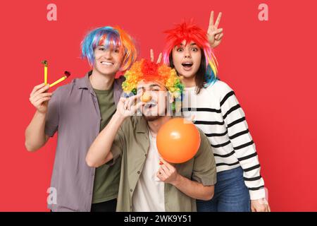 Junge Freunde in lustiger Verkleidung mit Partypfeife und Ballon auf rotem Hintergrund. Aprilfeier zum Narrentag Stockfoto