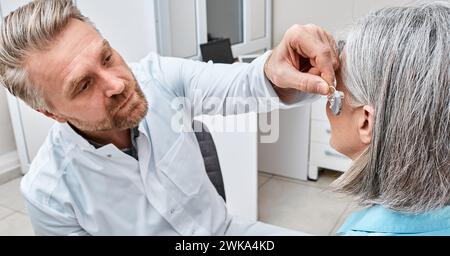 Audiologe, der ein Hörgerät am Ohr einer Frau mittleren Alters in einer Hörklinik installiert. Schwerhörigkeit, Hörlösungen Stockfoto