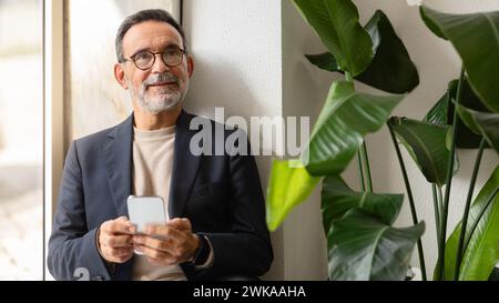 Nachdenklicher Seniorenmann in Blazer und Rollkragen, der ein Smartphone hält Stockfoto