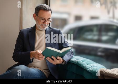 Ein nachdenklicher älterer Mann mit Brille, der in das Lesen eines Buches vertieft ist, bequem sitzend Stockfoto