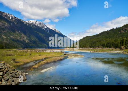 Blick auf einen Fluss und die fernen, schneebedeckten Berge an einem Frühlingstag, mit den Wolken, die sich im Wasser spiegeln. Stockfoto