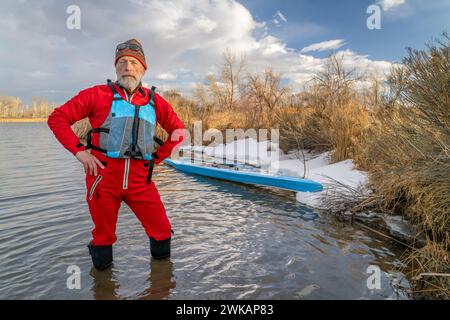 Der ältere Paddler mit Schwimmweste und Trockenanzug steht im Winter oder Frühjahr in Colorado neben seinem Stand Up Paddleboard auf dem See Stockfoto