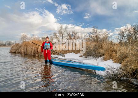 Der ältere Paddler mit Schwimmweste und Trockenanzug steht im Winter oder Frühjahr in Colorado neben seinem Stand Up Paddleboard auf dem See Stockfoto