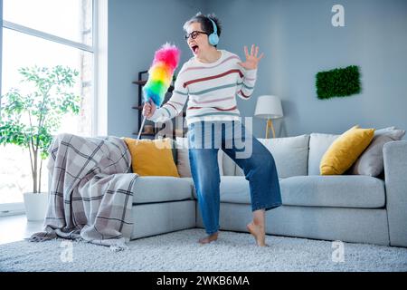 Ganzkörperporträt einer kühlen alten Dame, die Duster Dance Singen Musik genießen Kopfhörer geräumiges, modernes Haus drinnen Stockfoto