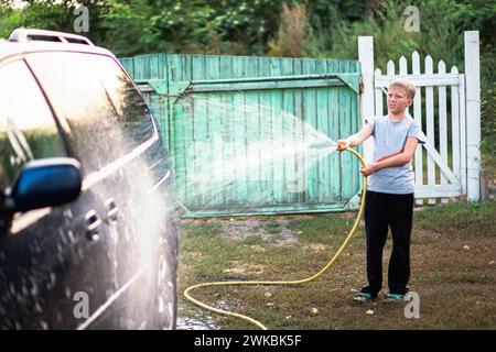 Ein Teenager wäscht ein Auto, indem er Wasser mit Wasser aus einem Gartenschlauch darauf gießt. Das Konzept der Familienzusammenarbeit Stockfoto