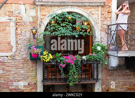 Fassade eines Gebäudes mit Blumen und Pflanzen und einem blonden Mädchen auf einem Balkon in Venedig, Italien Stockfoto