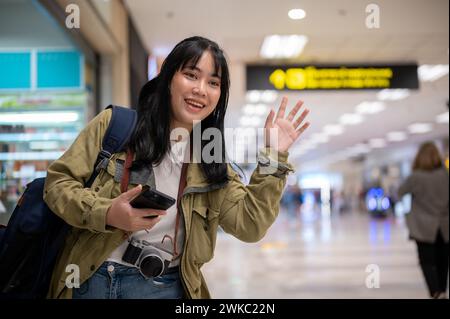 Eine fröhliche junge asiatische Reisende winkt mit der Hand, ruft ihre Freundin am Flughafen an und begrüßt sie. Stockfoto