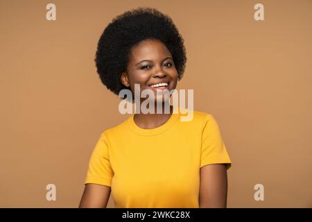 Fröhliche, offen lächelnde schwarze Frau, die in die Kamera blickt, isoliert in gelbem Hemd auf beigem Hintergrund Stockfoto
