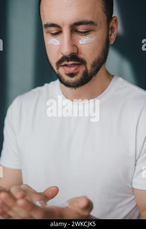 Porträt eines jungen Mannes, der auf dem Sofa sitzt, das Kosmetikprodukt hält und es auf das Gesicht aufträgt. Das Konzept der liebevollen Hautcreme. Stockfoto