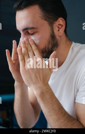 Seitenporträt eines jungen bärtigen männlichen Schmiercreme unter den Augen während der Hautpflege am Morgen. Stockfoto