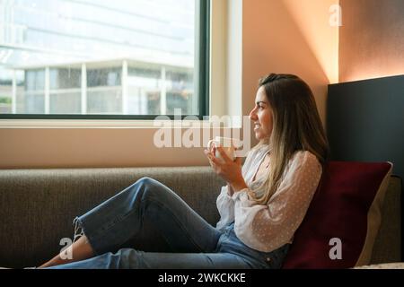 Eine junge Frau sitzt gemütlich am Fenster eines Hotelzimmers und genießt eine warme Tasse Kaffee, während sie nach draußen blickt und das einfache Vergnügen hervorhebt Stockfoto