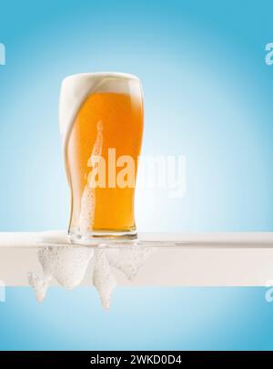 Glas blondes Bier mit Schaum auf weißem Regal auf blauem Hintergrund. Stockfoto