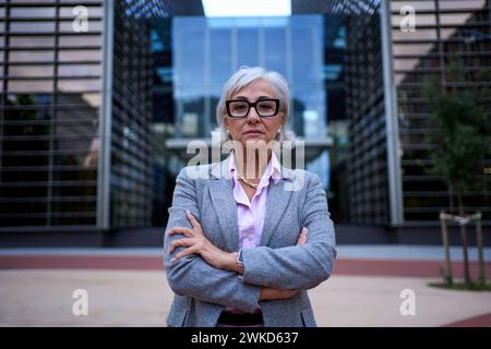 Erfolgreiche und ernsthafte reife Geschäftsfrau mit gekreuzten Armen am Arbeitsplatz und Blick in die Kamera. Stockfoto