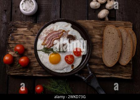 Spiegeleier mit Speck und Gemüse in gusseiserner Pfanne, auf alten dunklen Holztischhintergrund, Stockfoto