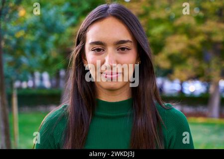 Porträt einer jungen kaukasischen Frau, die lächelnd in die Kamera schaut, draußen in einem öffentlichen Park steht. Stockfoto