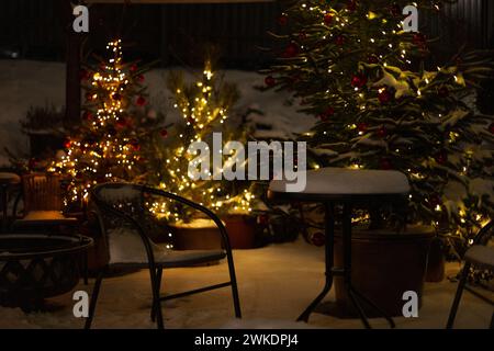 Wunderschöner Blick auf den Winter mit Weihnachtsbäumen, Schnee, Lichtern, Stuhl und Tisch auf der Terrasse in der Winternacht. Vintage Stockfoto