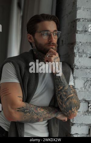 Porträt Eines gutaussehenden Hipster-Geschäftsmannes mit Bart und Frisur mit Tattoos mit Brille in Einem weißen T-Shirt mit Pullover Sta Stockfoto