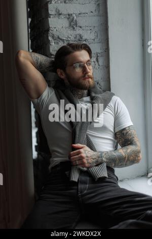 Stilvoller erfolgreicher männlicher Hipster-Geschäftsmann in Einem Bart mit Einem Tattoo mit Brille in Einem modischen weißen Hemd mit Einem Pullover sitzt neben dem Fenster Stockfoto