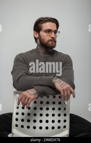 Der coole hübsche junge Hipster-Mann mit Frisur und Bart mit Brille in Einem modischen Strickpullover sitzt auf Einem weißen Stuhl im Studio Stockfoto