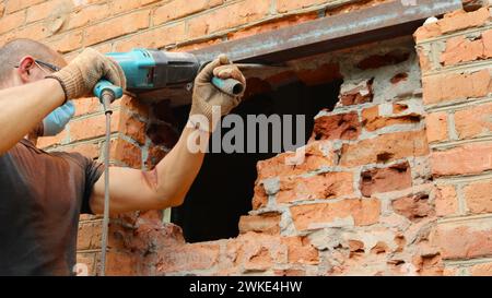 Ein Mann in Maske und Brille benutzt einen pneumatischen Bohrer, um eine Fensteröffnung in einer Ziegelmauer von der Straßenseite des Gebäudes auszuhöhlen Stockfoto