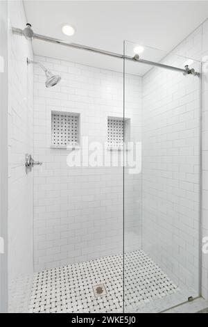 Eine begehbare Dusche mit weißen U-Bahn-Fliesen, Glasschiebetür, Chrom-Duschkopf und zwei Nischen-Regale mit Marmorfliesen. Stockfoto