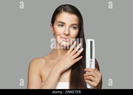 Attraktives Modell Frau hält Haareisen und glättet ihr gesundes langes dunkles Haar auf weißem Hintergrund. Haarpflege- und Haarstyling-Konzept Stockfoto