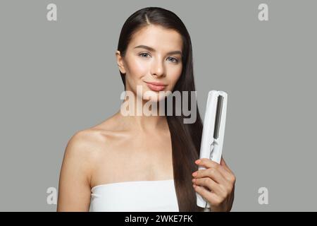 Hübsches Modell Frau hält Haareisen und glättet ihr gesundes langes dunkles Haar auf weißem Hintergrund. Haarpflege- und Haarstyling-Konzept Stockfoto