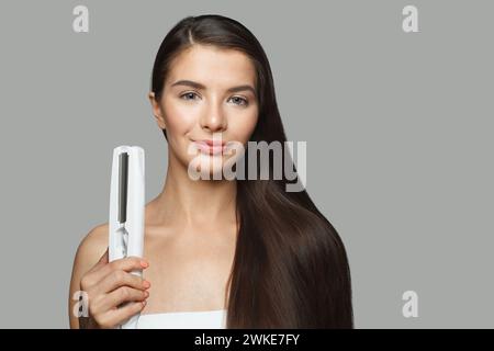 Schönes Modell Frau hält Haareisen und glättet ihr gesundes langes dunkles Haar auf weißem Hintergrund. Haarpflege- und Haarstyling-Konzept Stockfoto