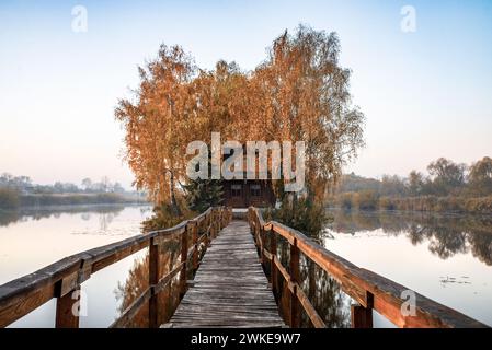 Morgenlandschaft eines kleinen Hauses auf einer künstlichen Insel mit einer langen Holzbrücke. Um das Haus herum ist ein kleiner See mit leichtem Nebel. Herbst ca. Stockfoto