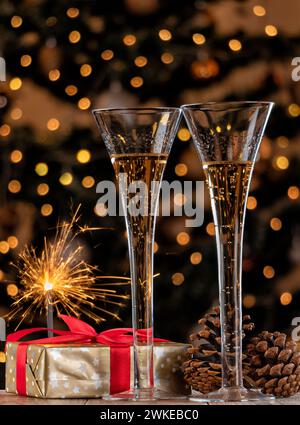 Zwei Gläser Champagner mit einem Weihnachtsgeschenk und brennenden Glitzern auf dem Hintergrund eines geschmückten Weihnachtsbaums. Stockfoto